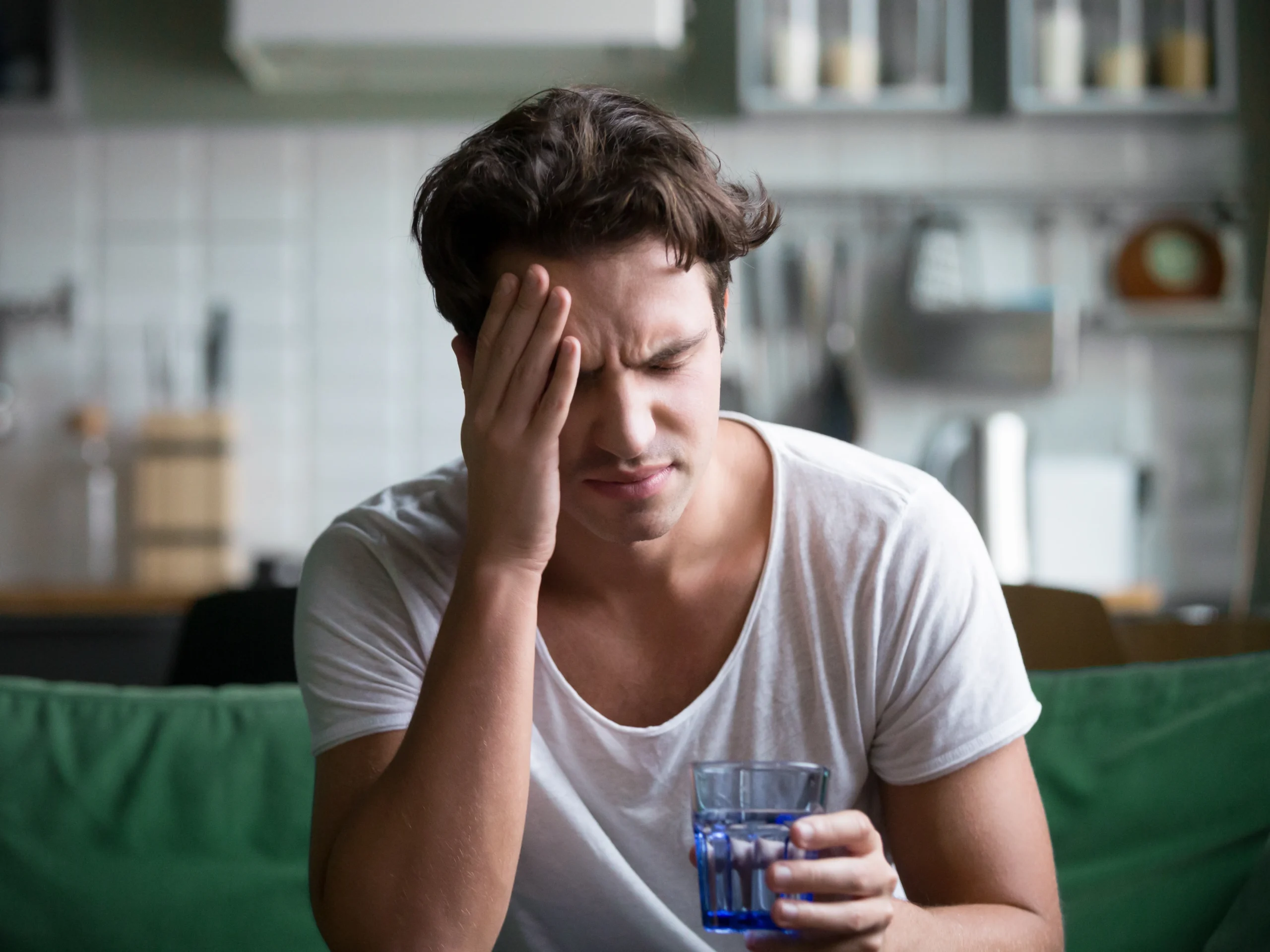 A young man with a severe headache or migraine sits with a glass of water.